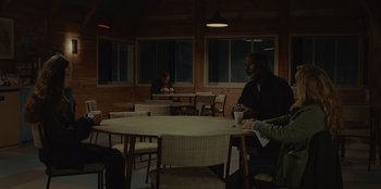 Movie still from “No Exit” (2022), directed by Damien Power – A group of people sitting at a table in a room; Wide shot, Over the shoulder angle