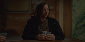 Movie still from “No Exit” (2022), directed by Damien Power – A man sitting at a table holding a deck of cards; Medium shot, Over the shoulder angle