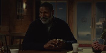 Movie still from “No Exit” (2022), directed by Damien Power – An older man sitting at a table with a cup of coffee in front of him; Medium shot, Over the shoulder angle