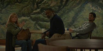 Movie still from “No Exit” (2022), directed by Damien Power – A man sitting in front of a painting of a mountain; Medium shot, High angle