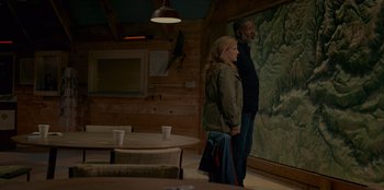 Movie still from “No Exit” (2022), directed by Damien Power – A man and a woman standing in front of a painting; Wide shot, High angle