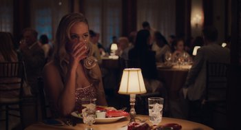 Movie still from “No Hard Feelings” (2023), directed by Gene Stupnitsky – A beautiful young lady sitting at a dinner table drinking champagne; Medium shot, Over the shoulder angle