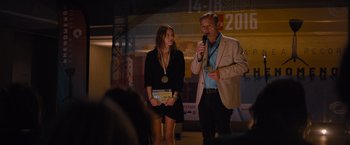 Movie still from “No Limit” (2022), directed by David M. Rosenthal – A man and a woman standing in front of a microphone; Medium shot, Over the shoulder angle