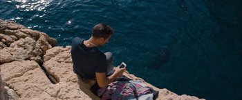Movie still from “No Limit” (2022), directed by David M. Rosenthal – A man sitting on a cliff looking at a phone; Wide shot, High angle