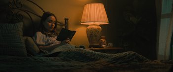 Movie still from “No One Will Save You” (2023), directed by Brian Duffield – A woman sitting in a bed reading a book next to a lamp; Medium shot, High angle