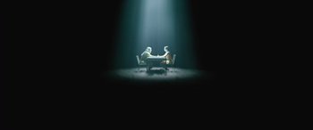 Movie still from “No One Will Save You” (2023), directed by Brian Duffield – Two people sitting at a table in the middle of a dark room; Extreme Wide shot, Overhead angle