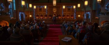 Movie still from “Noelle” (2019), directed by Marc Lawrence – An audience is sitting in a room with a man on a stage; Extreme Wide shot, High angle