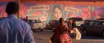 Movie still from “Noelle” (2019), directed by Marc Lawrence – A woman in a colorful dress walking in a parking lot near parked cars; Extreme Wide shot, Over the shoulder angle