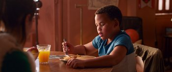 Movie still from “Noelle” (2019), directed by Marc Lawrence – A young boy sitting at a wooden table with a plate of food in front of him; Close Up shot, Over the shoulder angle