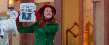 Movie still from “Noelle” (2019), directed by Marc Lawrence – A woman in a green coat and red hat holding a newspaper; Medium shot, Low angle