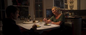 Movie still from “Notorious Nick” (2021), directed by Aaron Leong – A woman sitting at a table with a plate of food; Medium shot, Over the shoulder angle