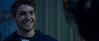 Movie still from “Notorious Nick” (2021), directed by Aaron Leong – A young man smiling for the camera in front of a building; Close Up shot, Over the shoulder angle