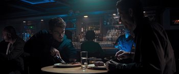 Movie still from “Notorious Nick” (2021), directed by Aaron Leong – Two men sitting at a table in front of plates of food; Medium shot, Over the shoulder angle