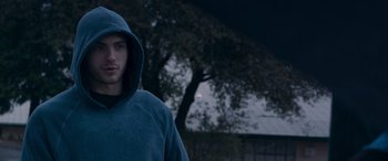 Movie still from “Notorious Nick” (2021), directed by Aaron Leong – A man wearing a hooded sweatshirt standing in front of a tree at night; Close Up shot, Low angle