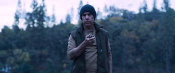 Movie still from “Notorious Nick” (2021), directed by Aaron Leong – A man holding a cell phone while standing in front of trees; Medium shot, Low angle