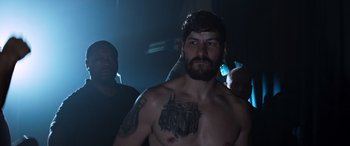 Movie still from “Notorious Nick” (2021), directed by Aaron Leong – A man with a beard and tattoos standing in a dark room; Medium shot, Low angle