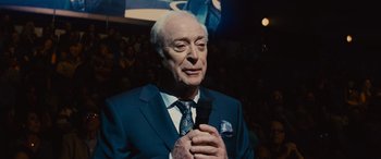 Movie still from “Now You See Me” (2013), directed by Louis Leterrier – An older man holding a microphone in front of an audience; Close Up shot, Over the shoulder angle