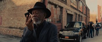 Movie still from “Now You See Me” (2013), directed by Louis Leterrier – An older man wearing a black hat smoking a cigarette while standing on the side of the road; Close Up shot, Low angle