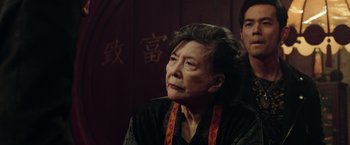 Movie still from “Now You See Me 2” (2016), directed by Jon M. Chu – An older woman wearing a black shirt and a red ribbon around her neck; Close Up shot, Over the shoulder angle