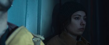 Movie still from “Nowhere” (2023), directed by Albert Pintó – A woman in a yellow jacket and a black hat is leaning against a wall; Close Up shot, Over the shoulder angle