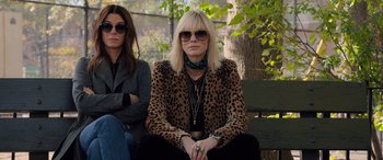 Movie still from “Ocean's Eight” (2018), directed by Gary Ross – Two women sitting on a bench wearing sunglasses; Medium shot, Over the shoulder angle