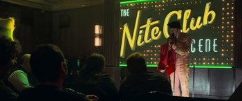 Movie still from “Ocean's Twelve” (2004), directed by Steven Soderbergh – A man standing in front of an audience in front of a lit up sign; Medium shot, Over the shoulder angle