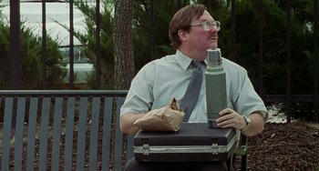 Movie still from “Office Space” (1999), directed by Mike Judge – A man sitting on a park bench with a lunch box and a bottle; Medium shot, Low angle