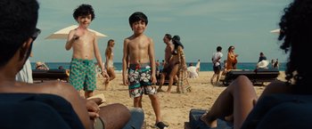 Movie still from “Old” (2021), directed by M. Night Shyamalan – A young boy standing on the beach near a crowd of people; Medium shot, Over the shoulder angle