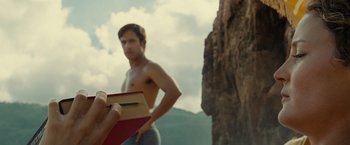 Movie still from “Old” (2021), directed by M. Night Shyamalan – A shirtless man reading a book near a cliff; Medium shot, Low angle