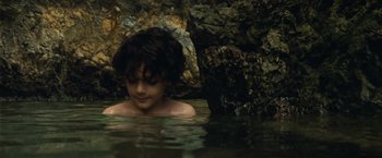 Movie still from “Old” (2021), directed by M. Night Shyamalan – A young boy is swimming in a body of water near rocks; Close Up shot, High angle