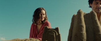 Movie still from “Old” (2021), directed by M. Night Shyamalan – A woman in a pink shirt is standing in front of a sand castle; Medium shot, Over the shoulder angle