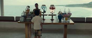 Movie still from “Old” (2021), directed by M. Night Shyamalan – Two people standing at a table with jars of candy; Wide shot, Over the shoulder angle