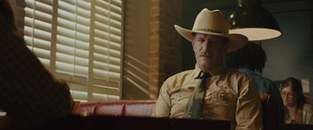 Movie still from “One Ranger” (2023), directed by Jesse V. Johnson – A man wearing a cowboy hat and a tie sitting on a red couch; Medium shot, Low angle