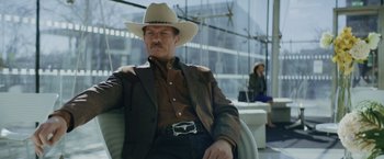 Movie still from “One Ranger” (2023), directed by Jesse V. Johnson – A man wearing a cowboy hat sitting in a chair; Medium shot, Low angle