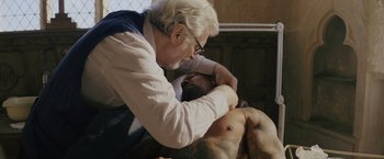 Movie still from “One Ranger” (2023), directed by Jesse V. Johnson – An older man is touching a shirtless man's chest; Medium shot, Over the shoulder angle