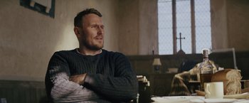 Movie still from “One Ranger” (2023), directed by Jesse V. Johnson – A man sitting at a table with his arms crossed; Medium shot, Over the shoulder angle