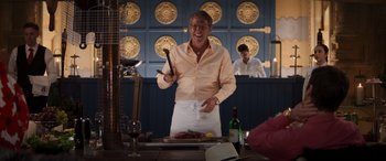 Movie still from “Operation Fortune: Ruse de Guerre” (2023), directed by Guy Ritchie – A man holding a knife in his hand while standing in front of a plate of food; Medium shot, Low angle