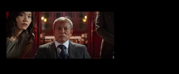 Movie still from “Operation Fortune: Ruse de Guerre” (2023), directed by Guy Ritchie – An older man wearing a suit and tie in front of a red door; Medium shot, Over the shoulder angle