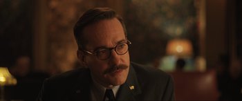 Movie still from “Operation Mincemeat” (2021), directed by John Madden – A person wearing glasses and a suit; Close Up shot, Over the shoulder angle