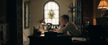Movie still from “Operation Mincemeat” (2021), directed by John Madden – A man sitting at a desk in front of a stained glass window; Medium shot, Low angle
