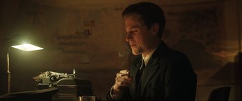 Movie still from “Operation Mincemeat” (2021), directed by John Madden – A man in a suit smoking a cigarette in a dimly - lit room; Medium shot, Low angle