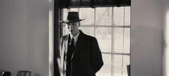 Movie still from “Oppenheimer” (2023), directed by Christopher Nolan – An older man wearing a suit and a hat standing in front of a window; Medium shot, Low angle