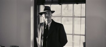 Movie still from “Oppenheimer” (2023), directed by Christopher Nolan – An older man wearing a suit and a hat standing in front of a window; Medium shot, Low angle
