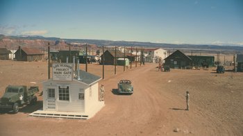 Movie still from “Oppenheimer” (2023), directed by Christopher Nolan – An old car parked in front of a building on the side of a dirt road; Extreme Wide shot, High angle