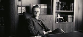 Movie still from “Oppenheimer” (2023), directed by Christopher Nolan – A man in a suit and tie sitting in an office chair; Medium shot, Low angle