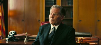 Movie still from “Oppenheimer” (2023), directed by Christopher Nolan – An older man wearing a suit and tie in an office setting; Close Up shot, Low angle
