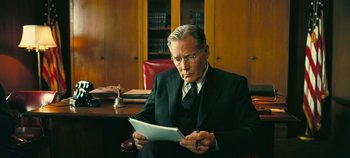Movie still from “Oppenheimer” (2023), directed by Christopher Nolan – An older man sitting at a desk in front of a book case; Medium shot, Low angle