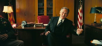 Movie still from “Oppenheimer” (2023), directed by Christopher Nolan – An older man in a suit sitting at a desk in front of a book case; Medium shot, Low angle