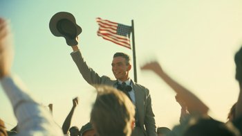 Movie still from “Oppenheimer” (2023), directed by Christopher Nolan – A man in a suit and hat raising his hat in the air in front of an american flag; Medium shot, Low angle
