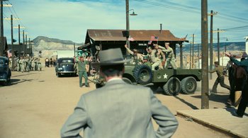 Movie still from “Oppenheimer” (2023), directed by Christopher Nolan – A man in a suit is riding on the back of an army truck; Wide shot, Over the shoulder angle